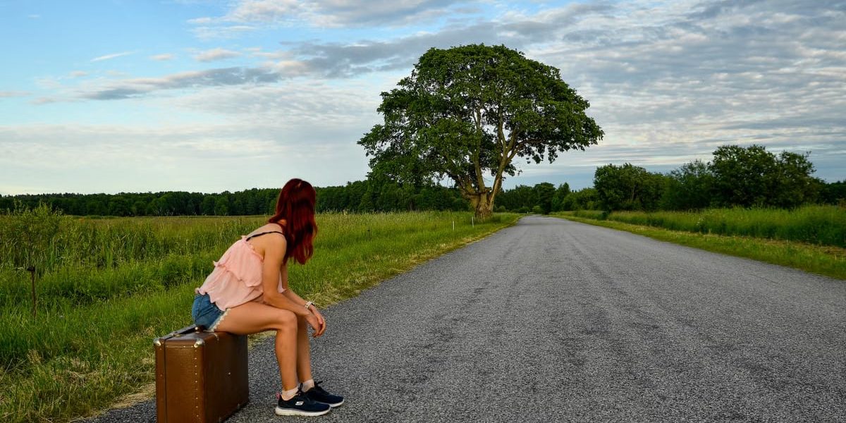 A woman in denim shorts and pink top sits on luggage waiting by the road in Estonia.