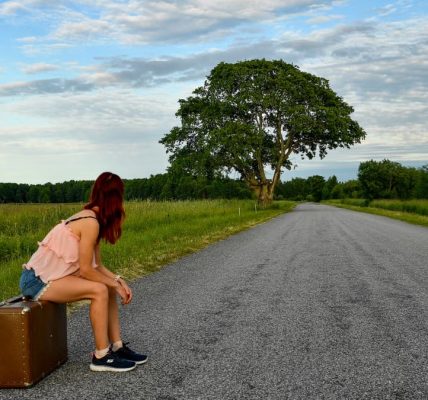 A woman in denim shorts and pink top sits on luggage waiting by the road in Estonia.