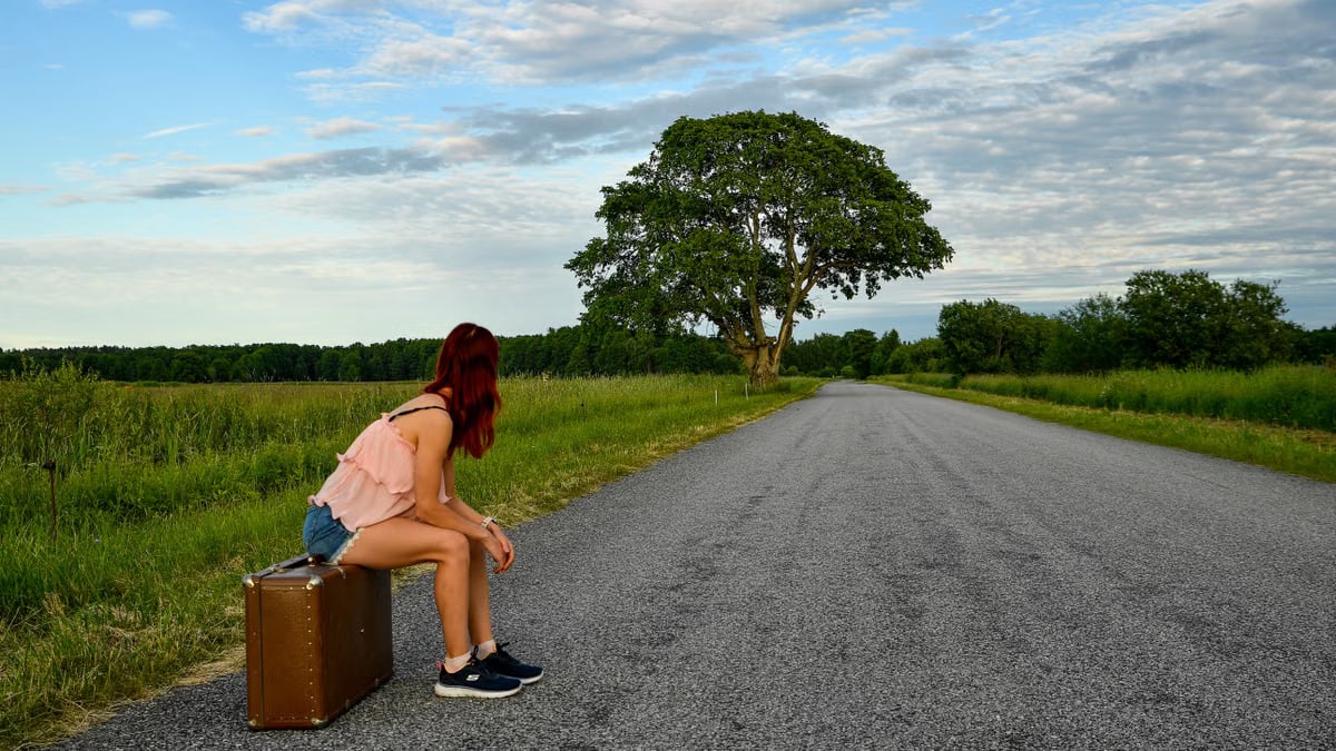 A woman in denim shorts and pink top sits on luggage waiting by the road in Estonia.